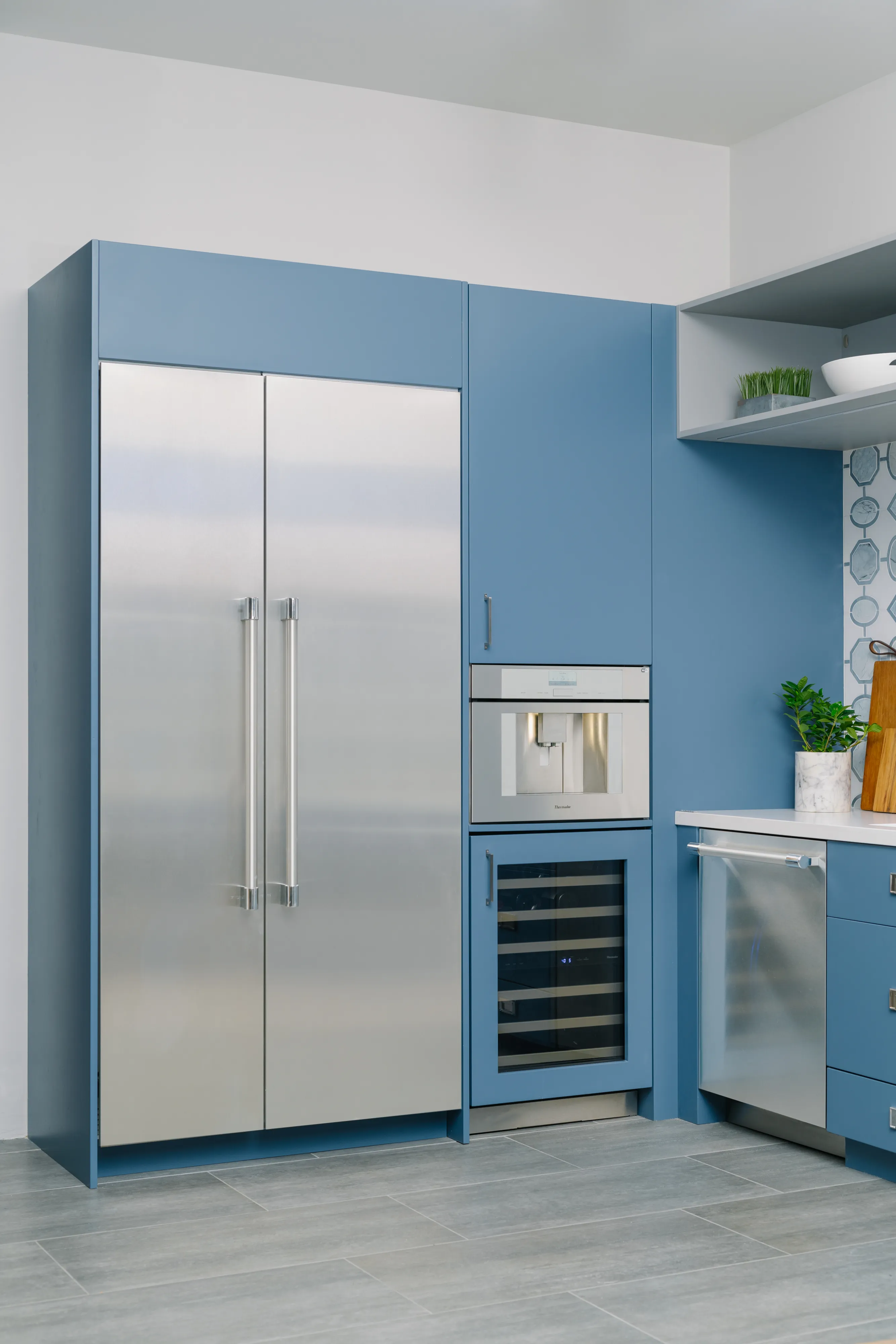 Contemporary kitchen scene with blue cabinetry: a stainless-steel side-by-side refrigerator with long vertical handles built into the blue cabinets, a built-in coffee dispenser above a glass-front wine cooler, a stainless-steel dishwasher to the right, a white countertop with a potted plant and cutting board, open shelving holding dishes, and gray tile floor.