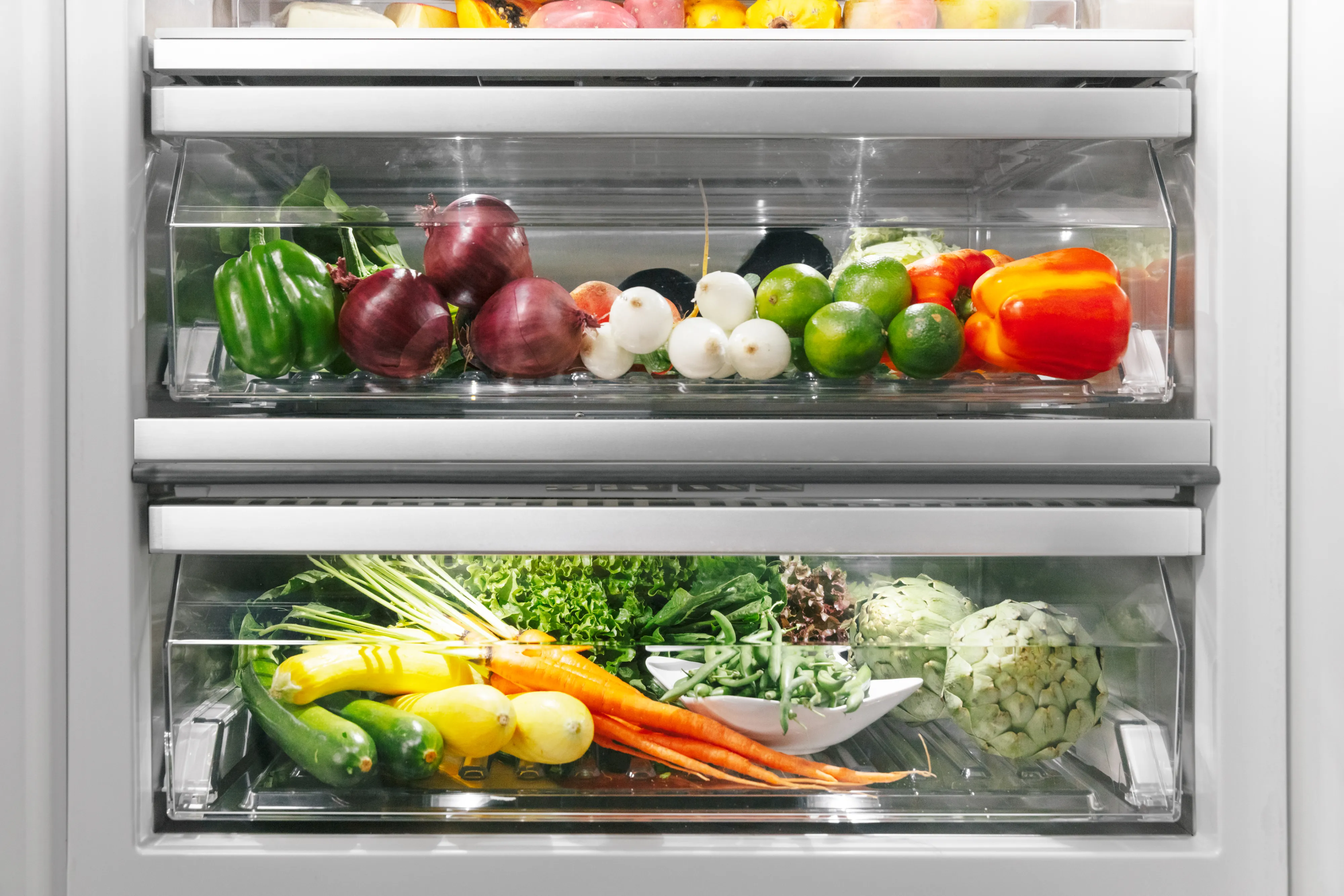 Close-up of two transparent refrigerator crisper drawers filled with fresh vegetables: the top drawer holds green bell peppers, red onions, small white radishes, limes and a red pepper; the bottom drawer contains carrots with tops, yellow squash/zucchini, assorted leafy greens and a head of cauliflower