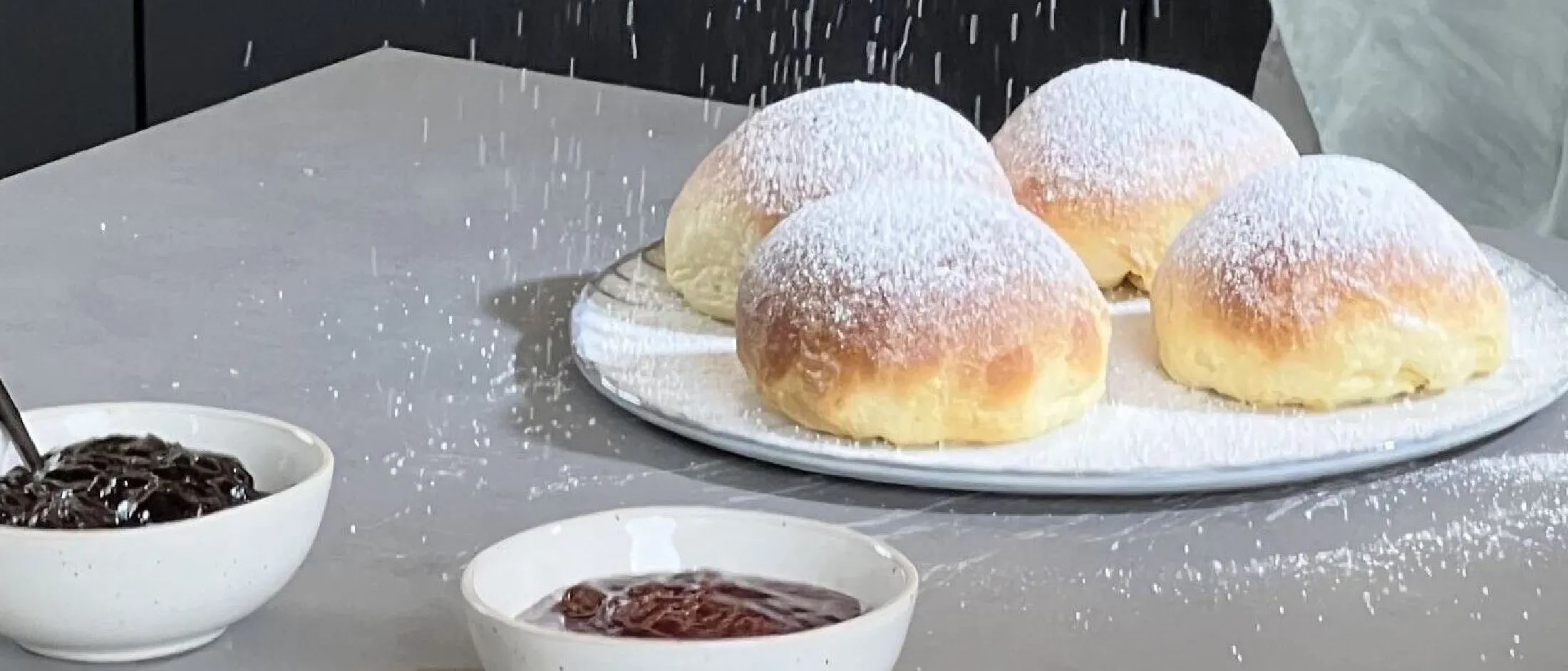 Vier mit Puderzucker bestäubte Krapfen auf einem weißen Teller neben zwei Schalen mit dunkler und roter Fruchtfüllung