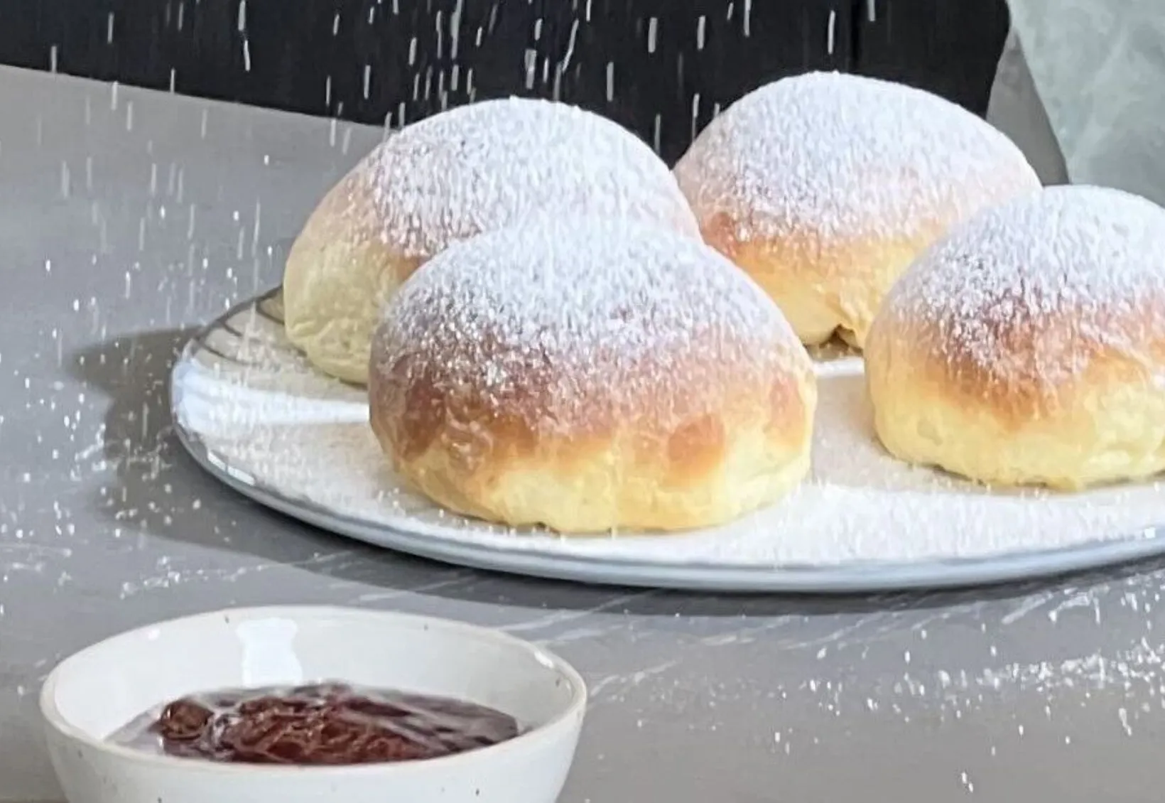 Vier mit Puderzucker bestäubte Krapfen auf einem weißen Teller neben zwei Schalen mit dunkler und roter Fruchtfüllung.