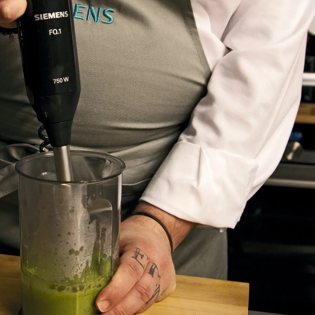 Chef utilizzando un frullatore ad immersione per purea di salsa verde in un contenitore chiaro su un bancone della cucina