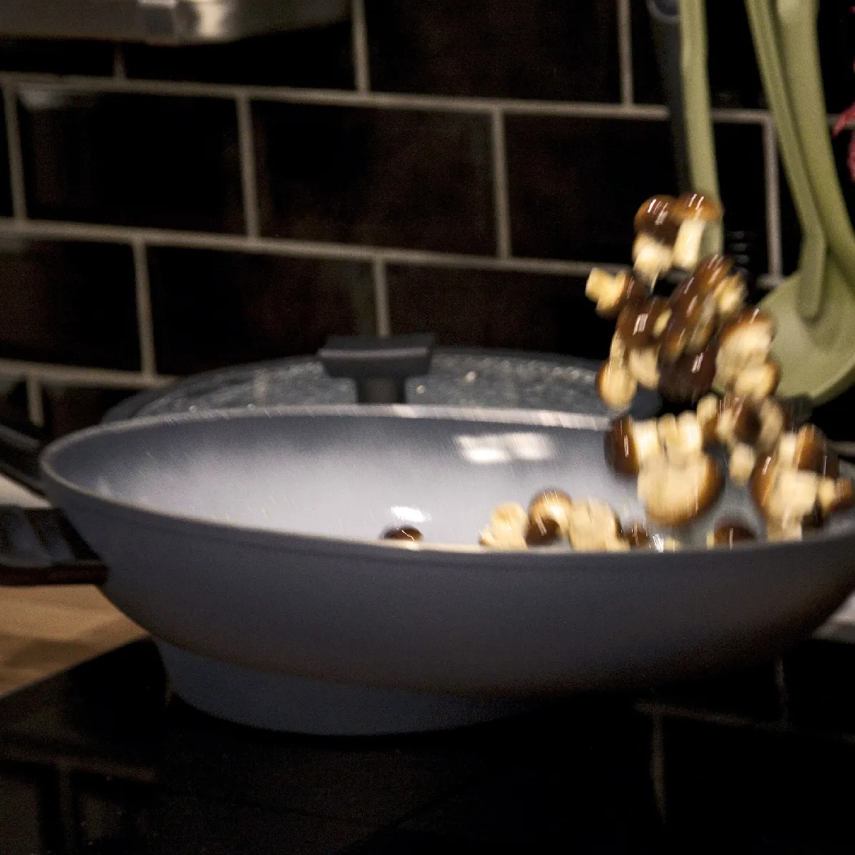 Funghi affettati che vengono capovolti in una padella grigia stile wok su un piano cottura, piastrelle nere backsplash