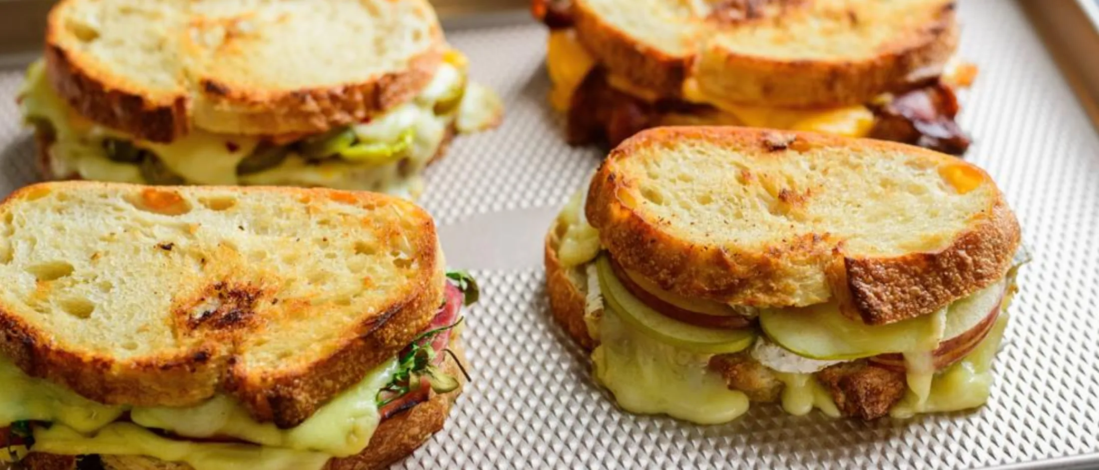 Four toasted grilled cheese sandwiches with melted cheese and sliced pickles on a baking sheetFour toasted grilled cheese sandwiches with melted cheese and sliced pickles on a baking sheet.