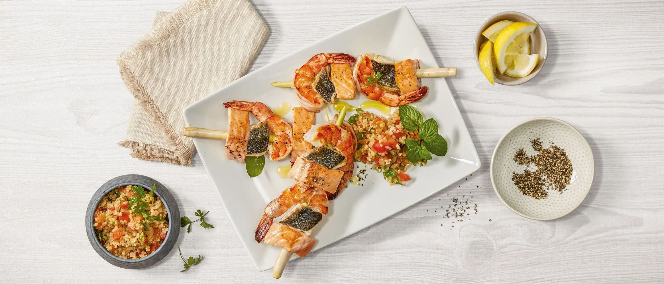 Spiedini di gamberi e salmone grigliati serviti con un contorno di insalata di bulgur, spicchi di limone e pepe nero su un piatto bianco.