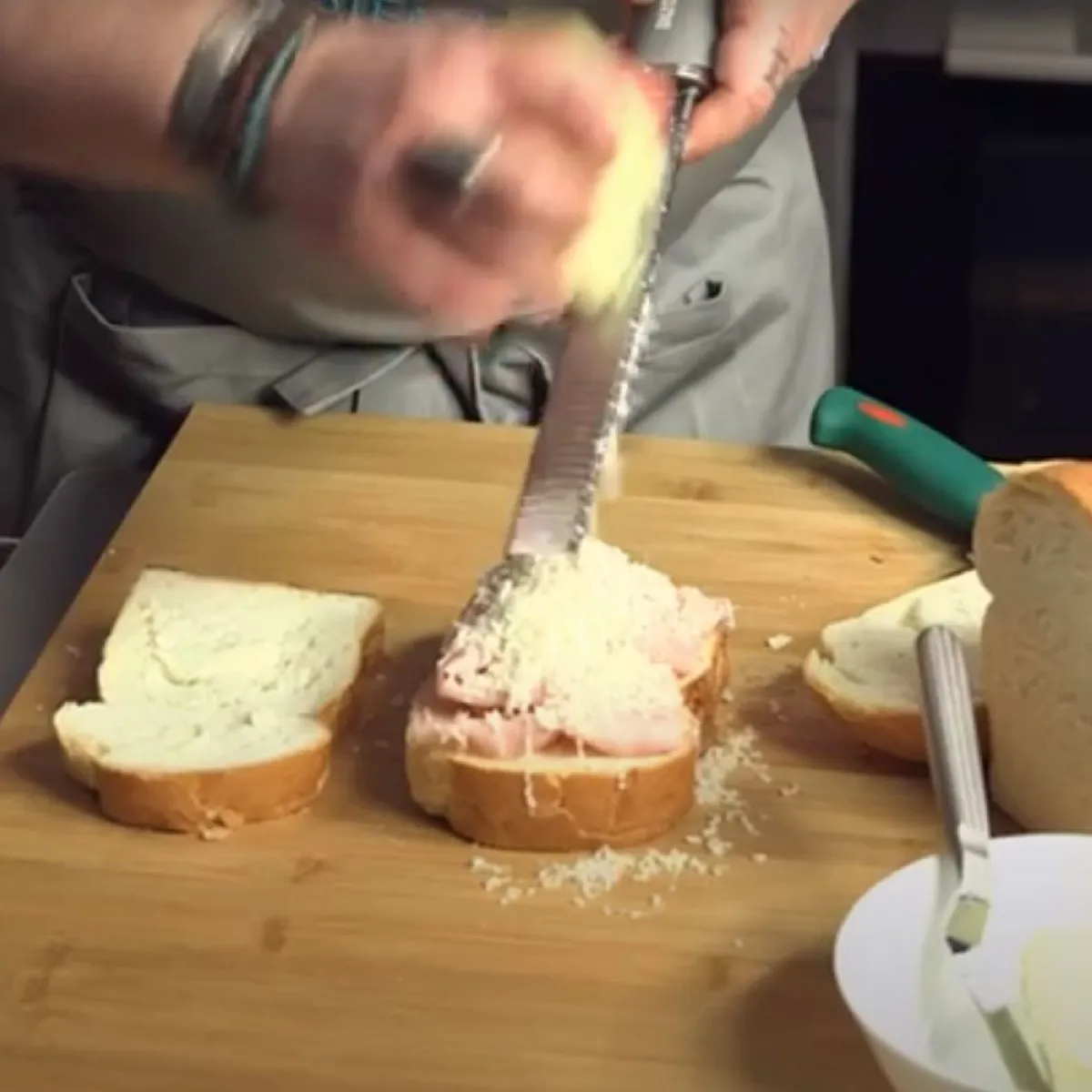 Mani che grattugiano formaggio su panini aperti su un tagliere di legno.