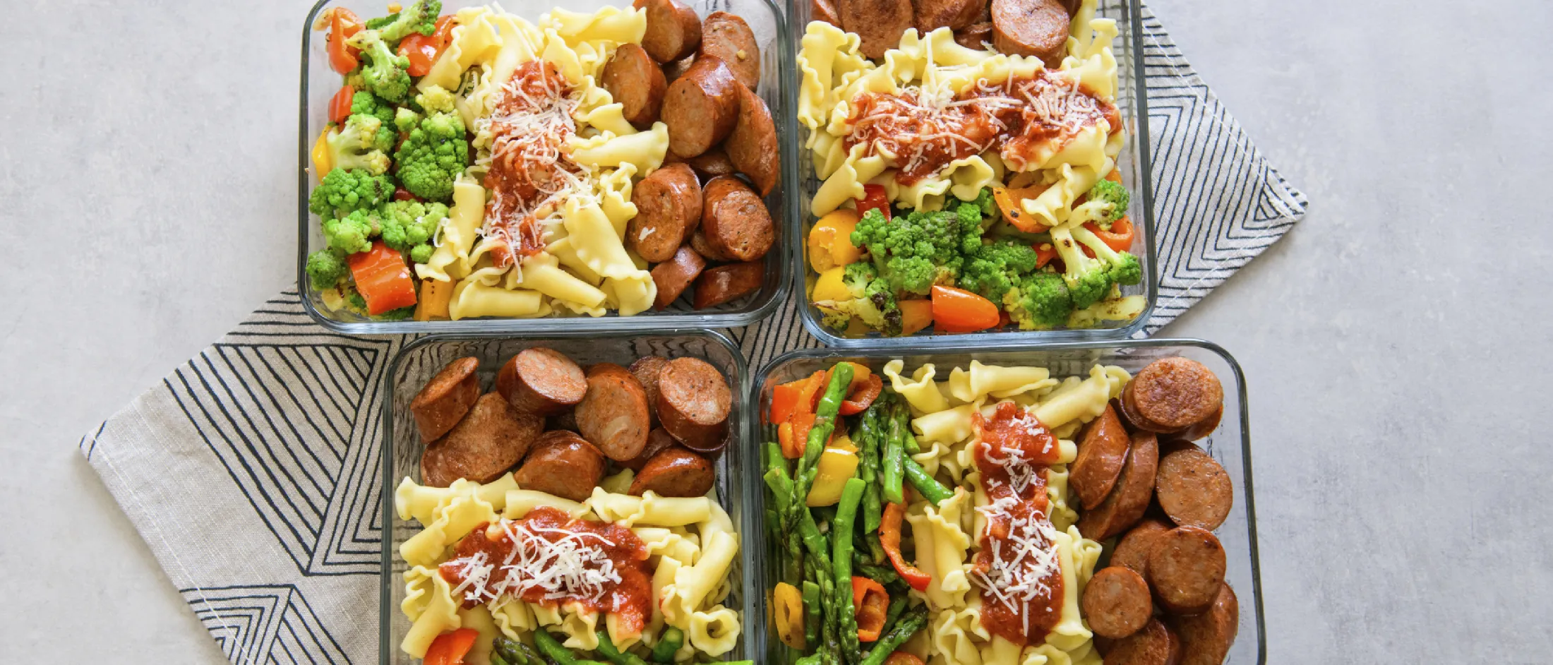 Four meal prep containers with pasta topped with tomato sauce and grated cheese, sliced sausage, and mixed steamed vegetables including broccoli, carrots, and asparagus