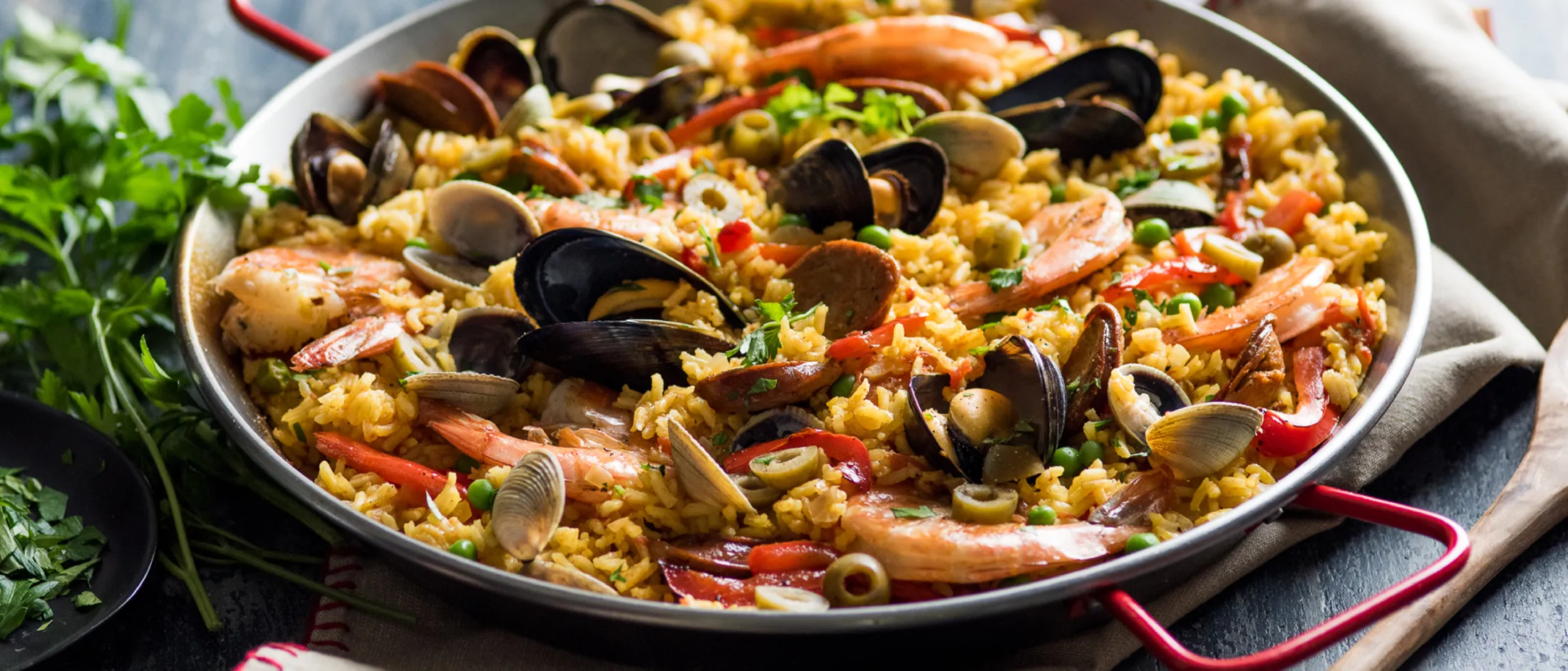 Pan of seafood paella with shrimp, mussels, clams, and rice on a cloth with herbs nearby