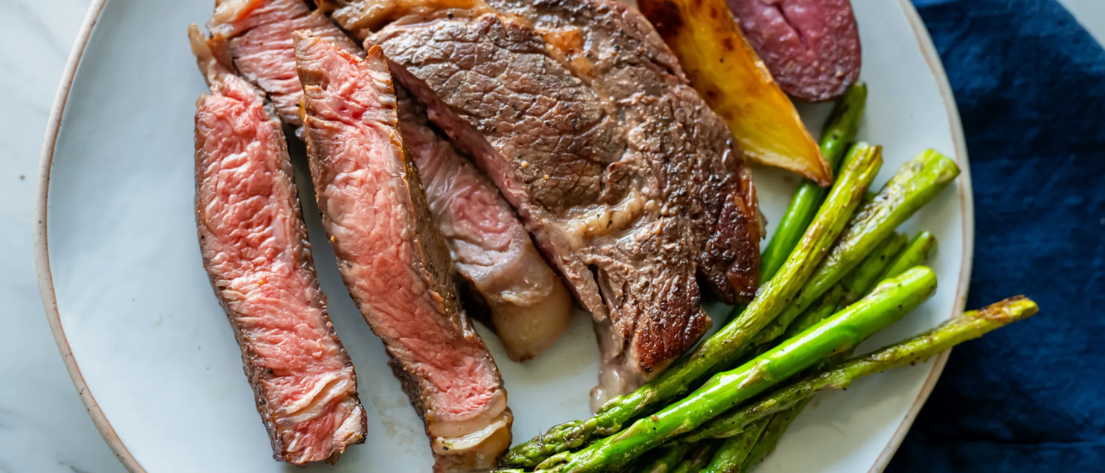 Plate with sliced medium-rare steak, roasted potato wedges, and grilled asparagus on a white marble surface with a blue napkin.