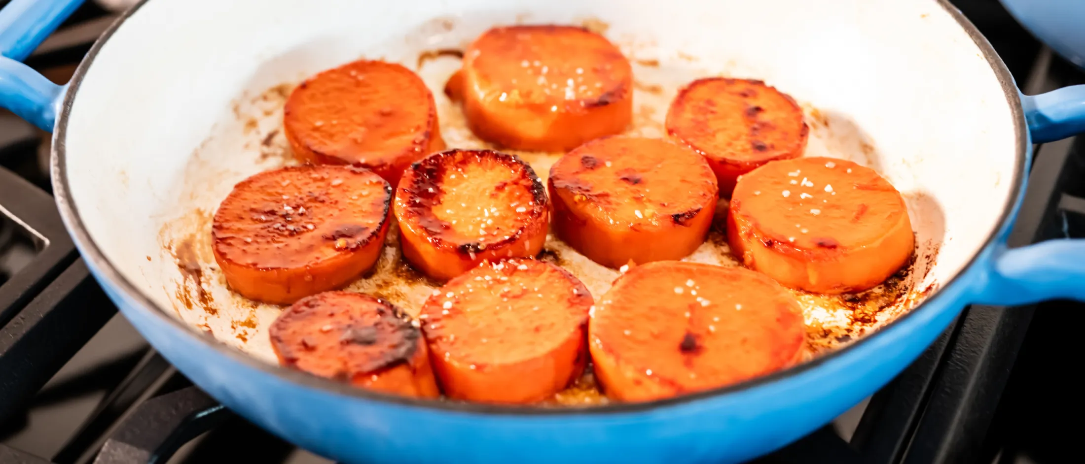 Slices of sweet potatoes cooking and caramelizing in a blue skillet on a gas stove burner.