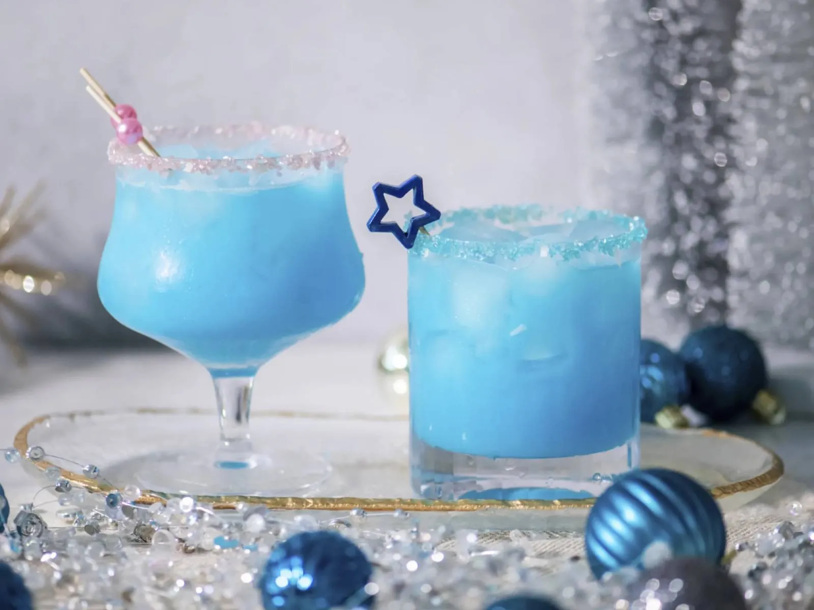 Two blue cocktails with salt rims on a tray, surrounded by silver and blue holiday decorations and beads.