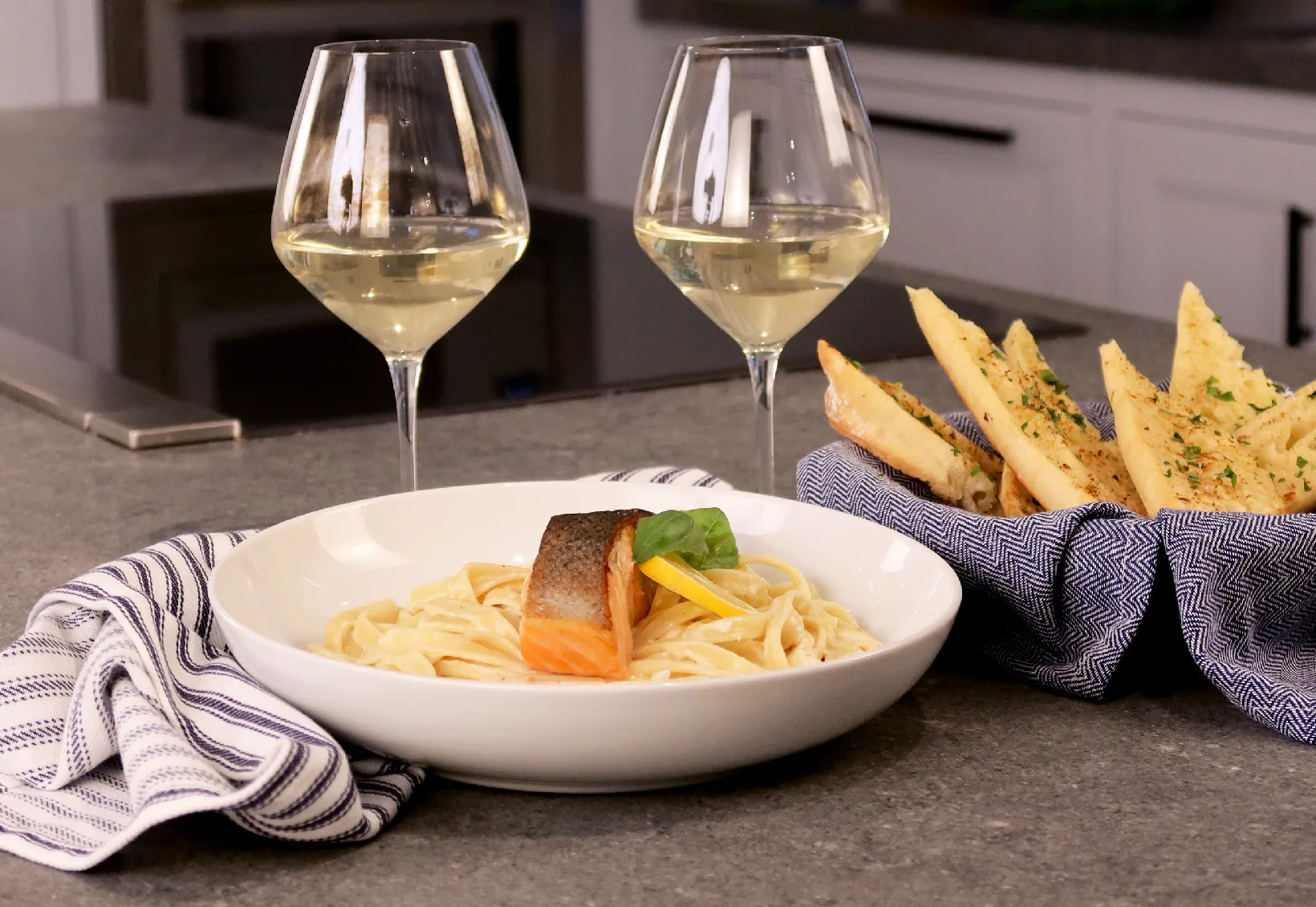 Plate with seared salmon on creamy pasta next to a basket of bread and two glasses of white wine on a kitchen counter