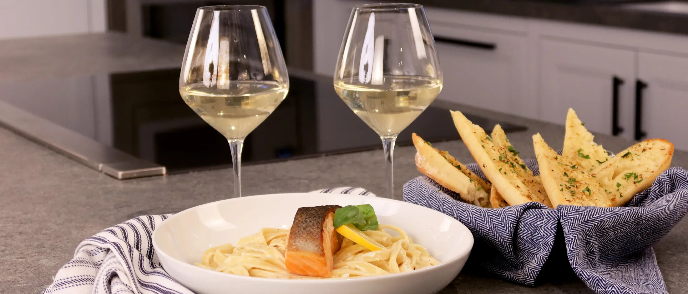 Plate with seared salmon on creamy pasta next to a basket of bread and two glasses of white wine on a kitchen counter