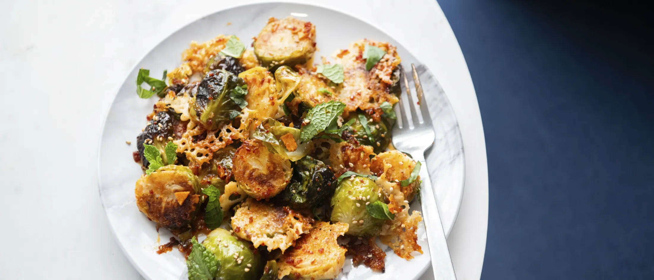 Plate of crispy roasted Brussels sprouts garnished with fresh herbs and sesame seeds, served with a fork on a white marble surface.