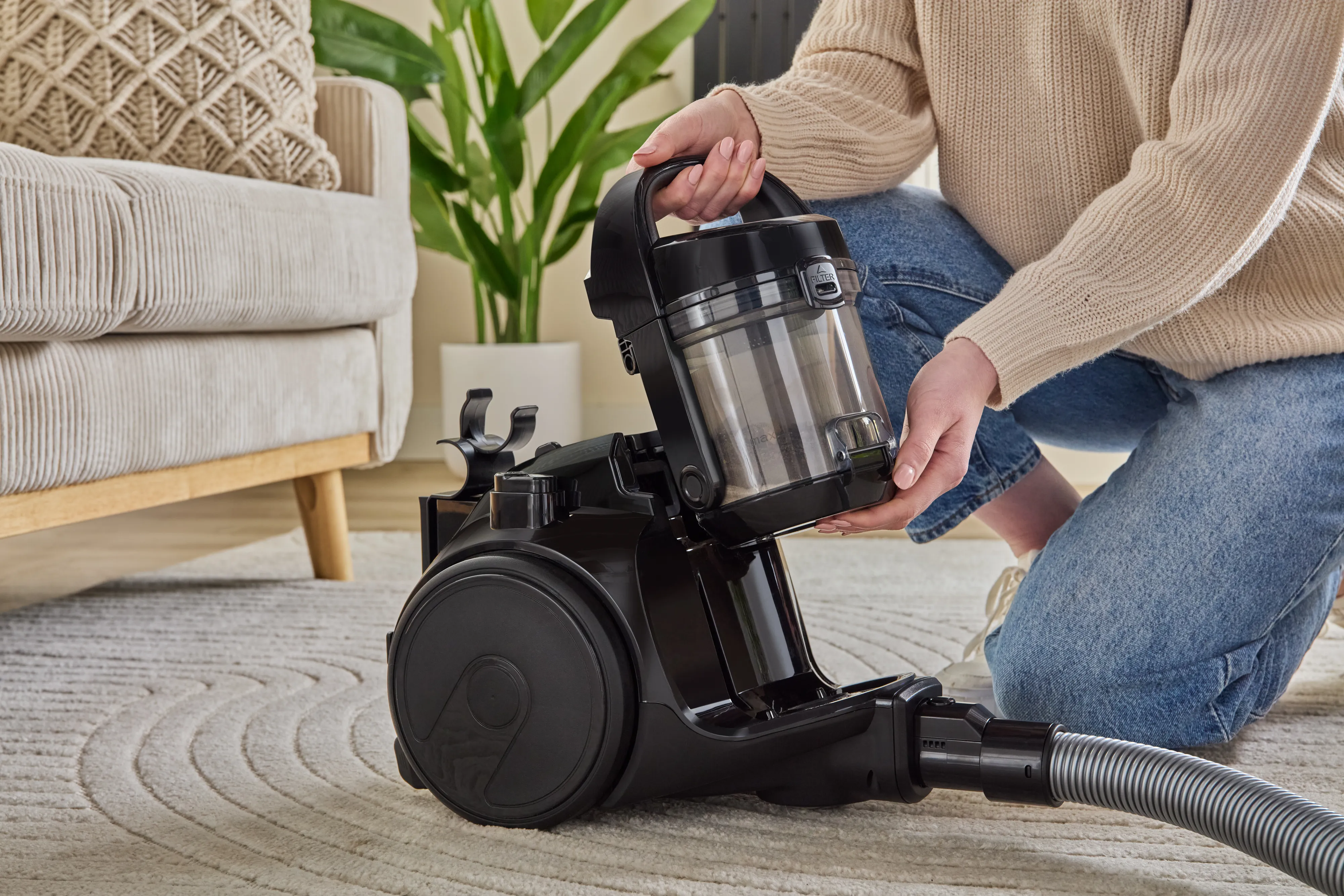 A woman empties the dustbin of a black Bosch bagless canister vacuum cleaner on a patterned rug in a living room.
