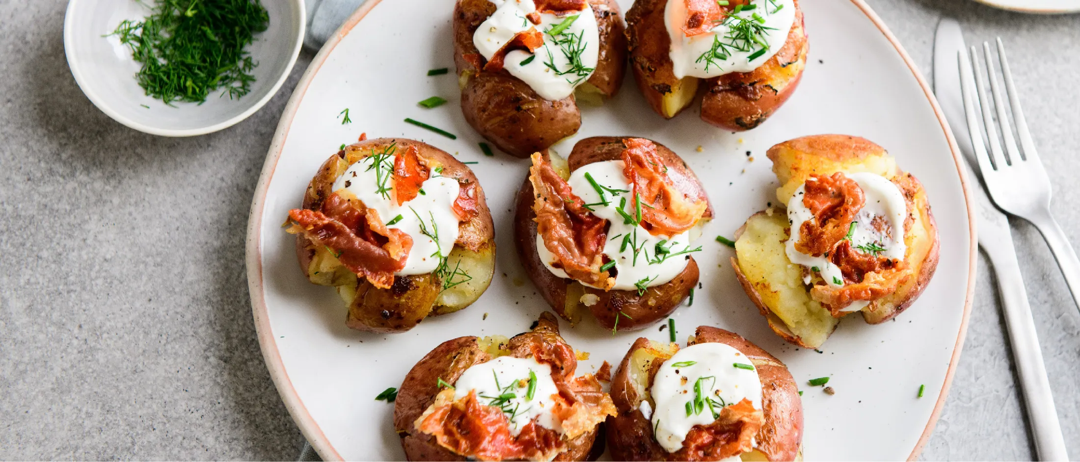 Plate with roasted red potatoes topped with sour cream, crispy prosciutto, and chopped herbs next to a small bowl of chopped herbs and a fork