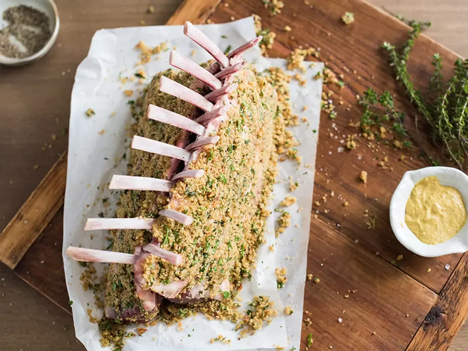 A rack of lamb, breaded with herbs and spices, rests on parchment paper on a wooden board. Mustard alongside.