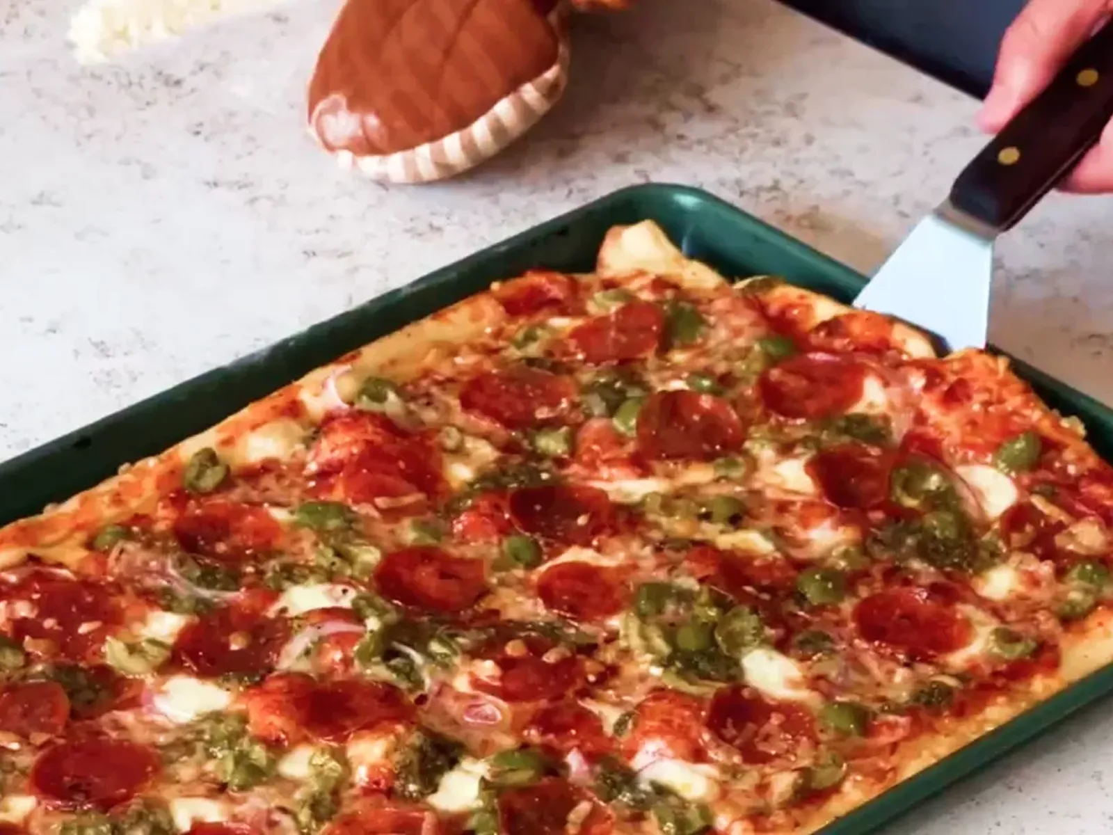 A metal spatula lifts a slice of rectangular pepperoni pizza with green peppers and red onions from a pan.