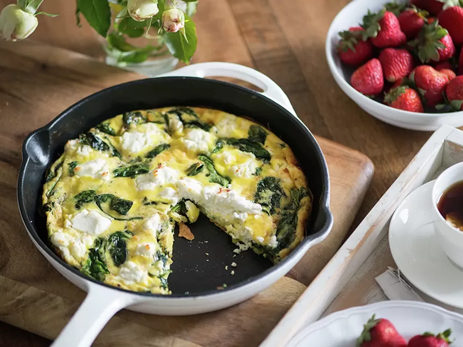 A cast-iron skillet holds a spinach and feta frittata with a slice removed, next to strawberries and tea.