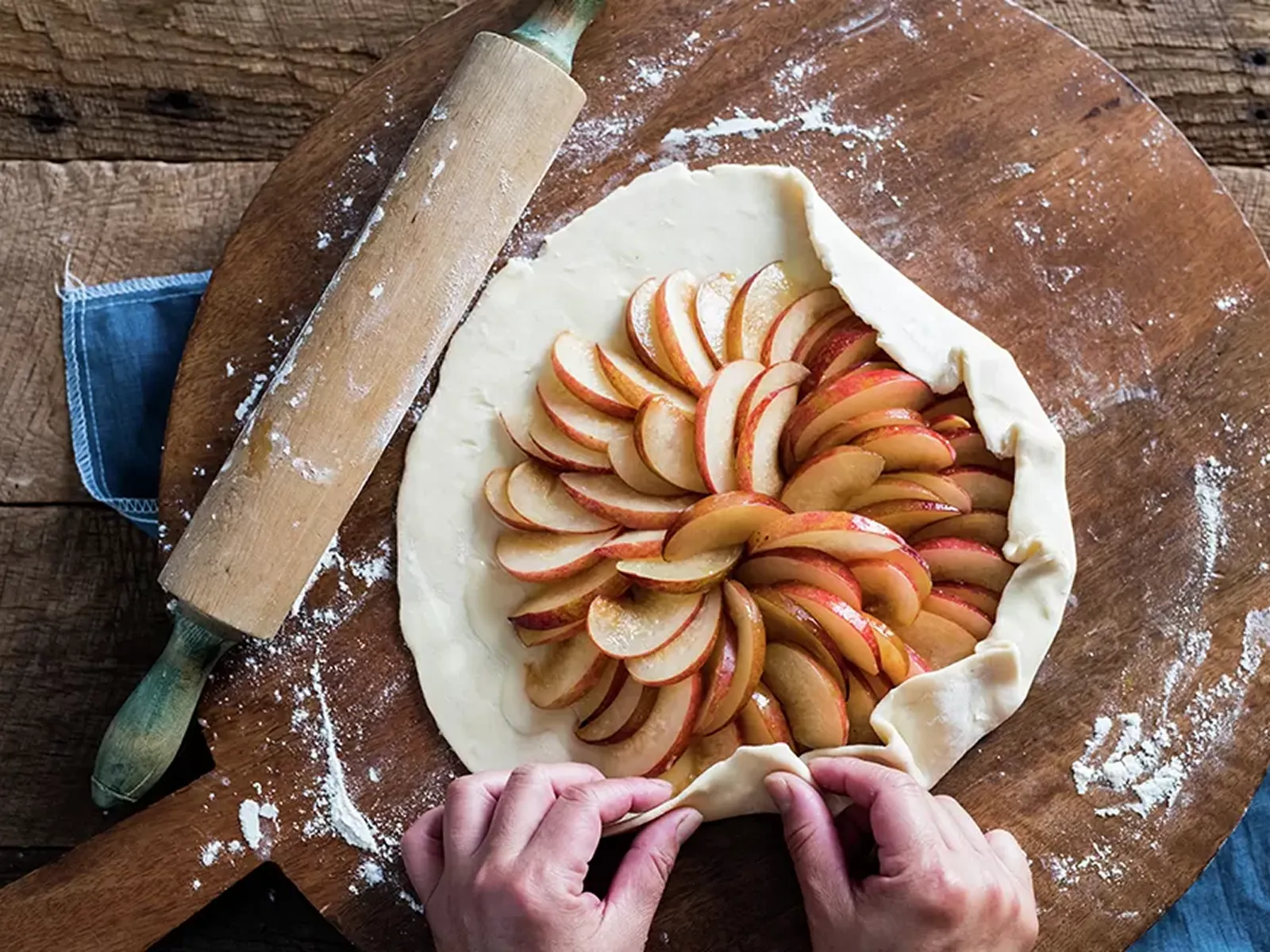 Hands arrange thinly sliced apples in a rustic tart on a wooden pizza peel, with a rolling pin nearby.