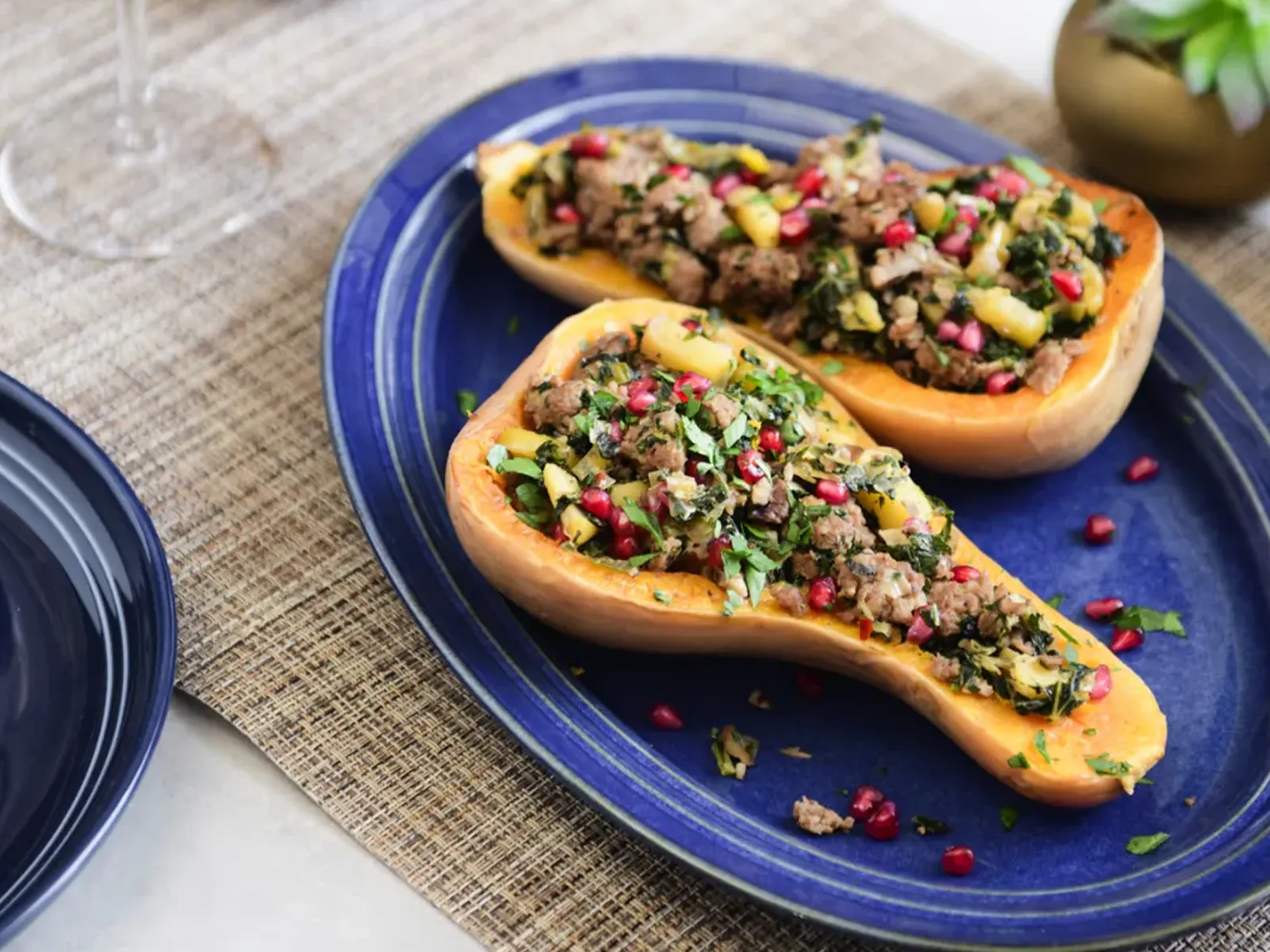 Two stuffed butternut squash halves, topped with ground meat, herbs, and pomegranate seeds, on a blue platter.