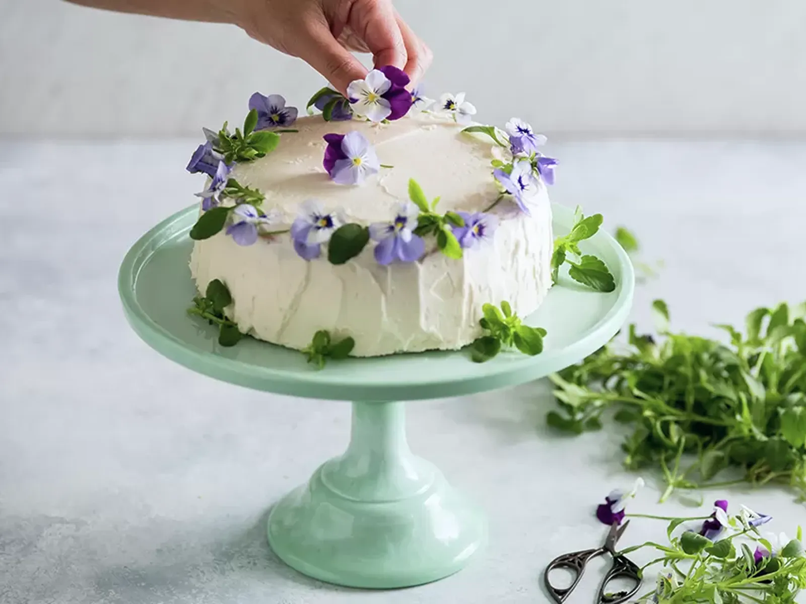 A hand delicately places purple and white edible flowers on top of a white frosted cake on a mint green stand.