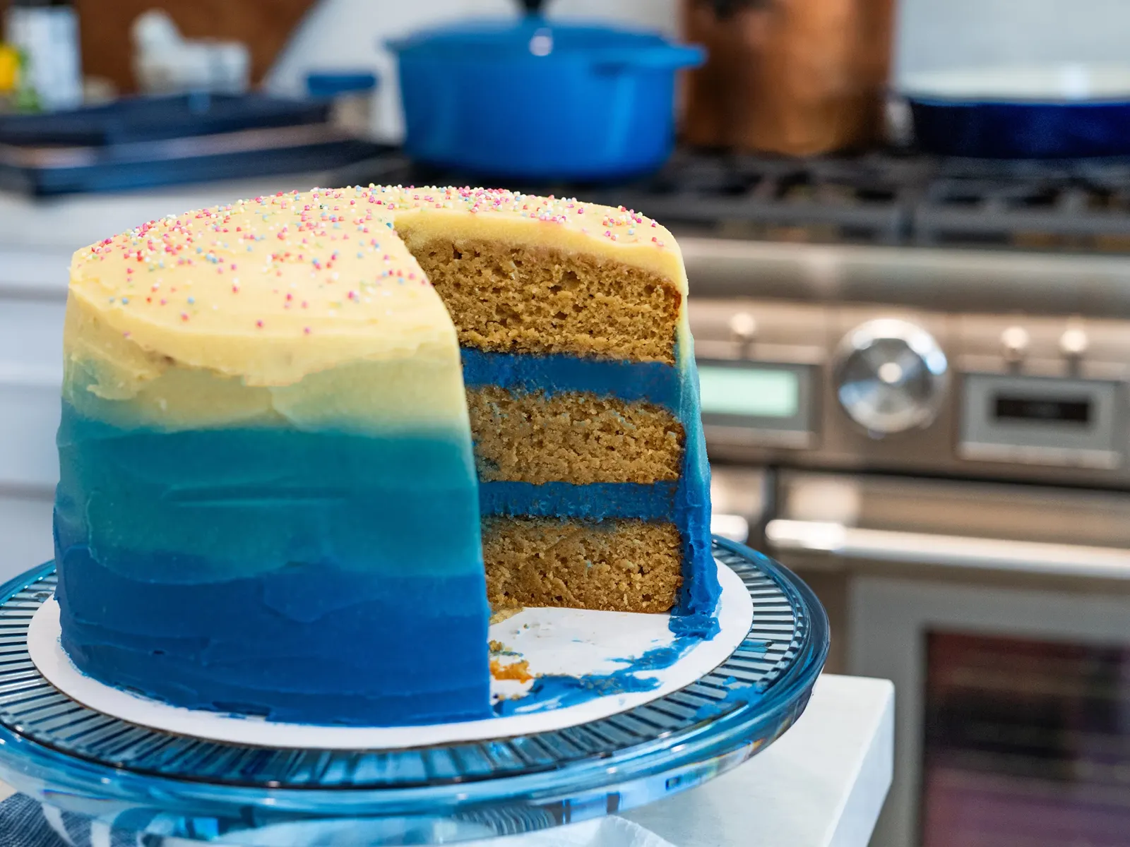 A slice of an ombre blue cake with brown layers and blue frosting, sitting on a clear cake stand.