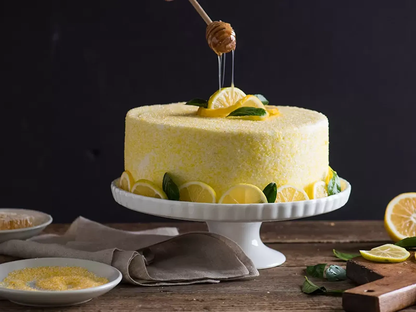 A beautiful lemon honey cake, covered in sugar, being drizzled with honey from a wooden dipper.