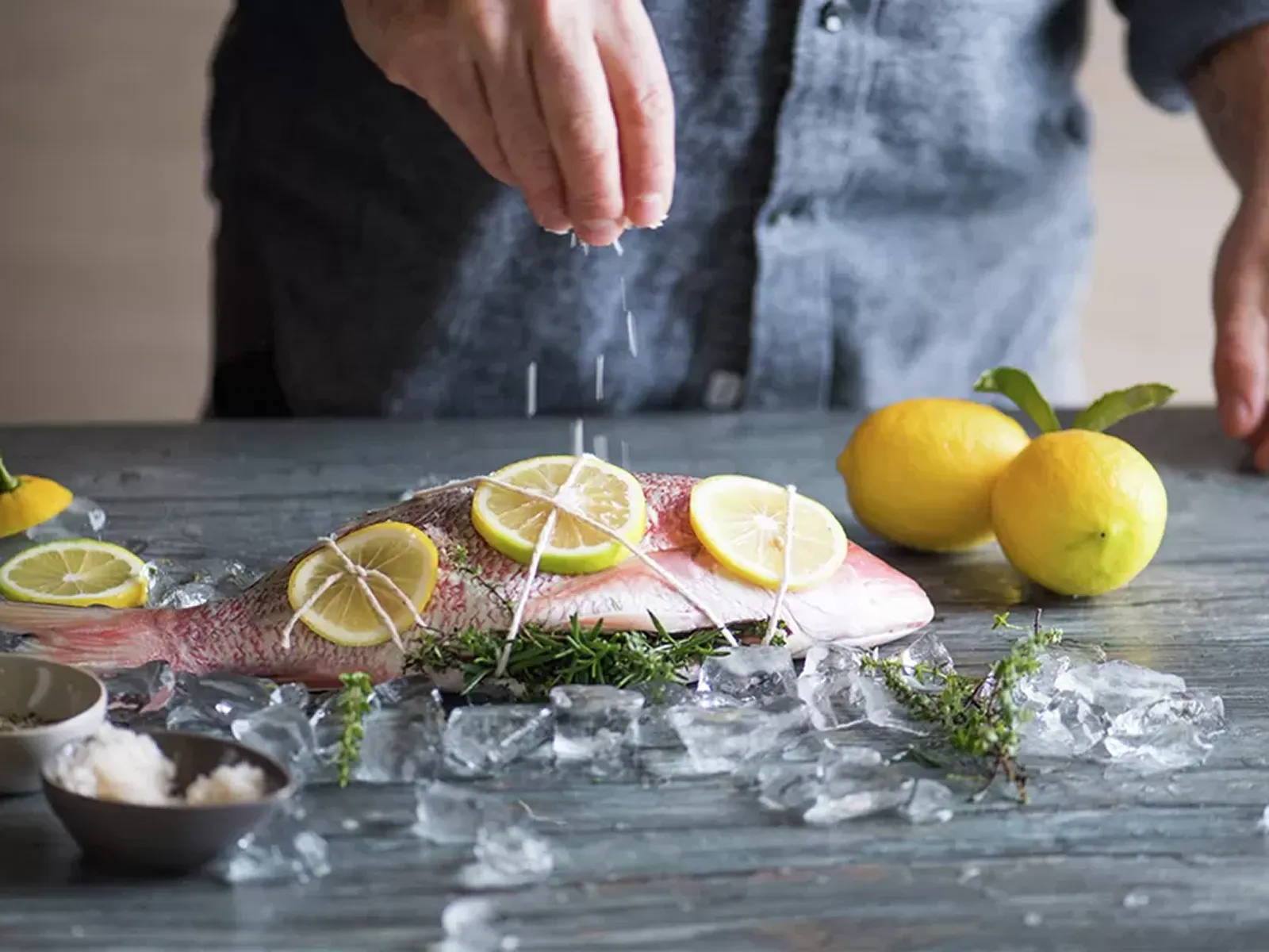 Hands sprinkle salt over a whole red snapper garnished with lemon and herbs, surrounded by ice and lemons.