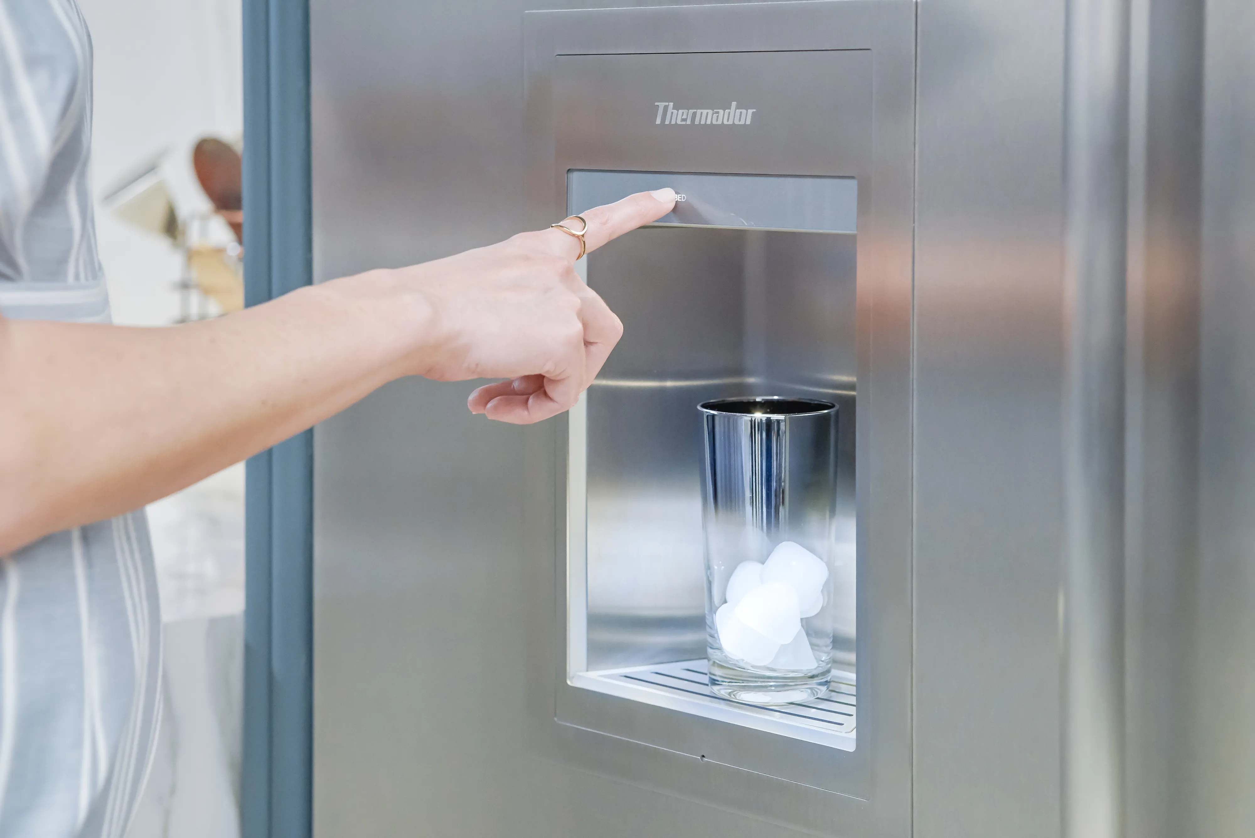Hand pressing button on a stainless steel refrigerator ice dispenser, dispensing ice cubes into a glass.