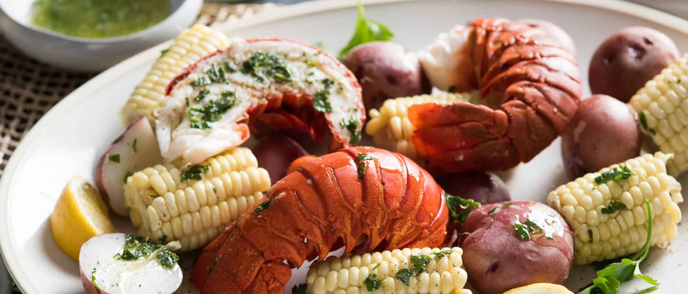 Plate with cooked lobster tails, corn on the cob, red potatoes, lemon wedges, and green herb garnish