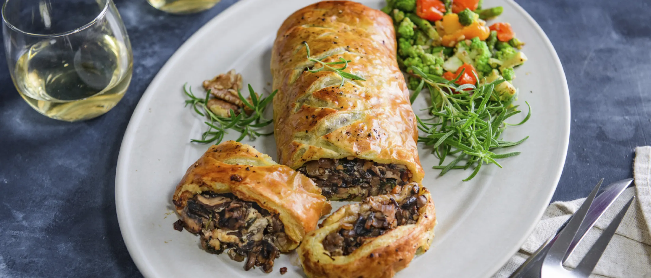 Plate with sliced mushroom Wellington, mixed steamed vegetables, fresh rosemary, walnuts, and two glasses of white wine on a dark surface.