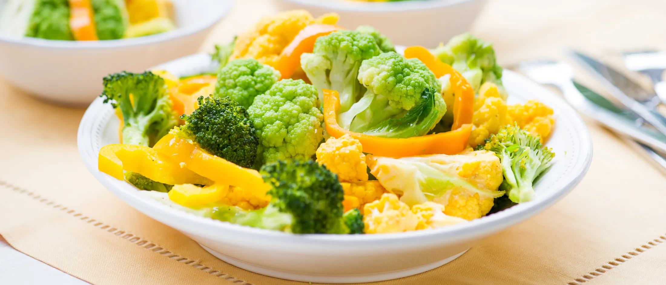 Three white bowls filled with steamed broccoli, yellow cauliflower, and yellow bell pepper slices on a light tablecloth.