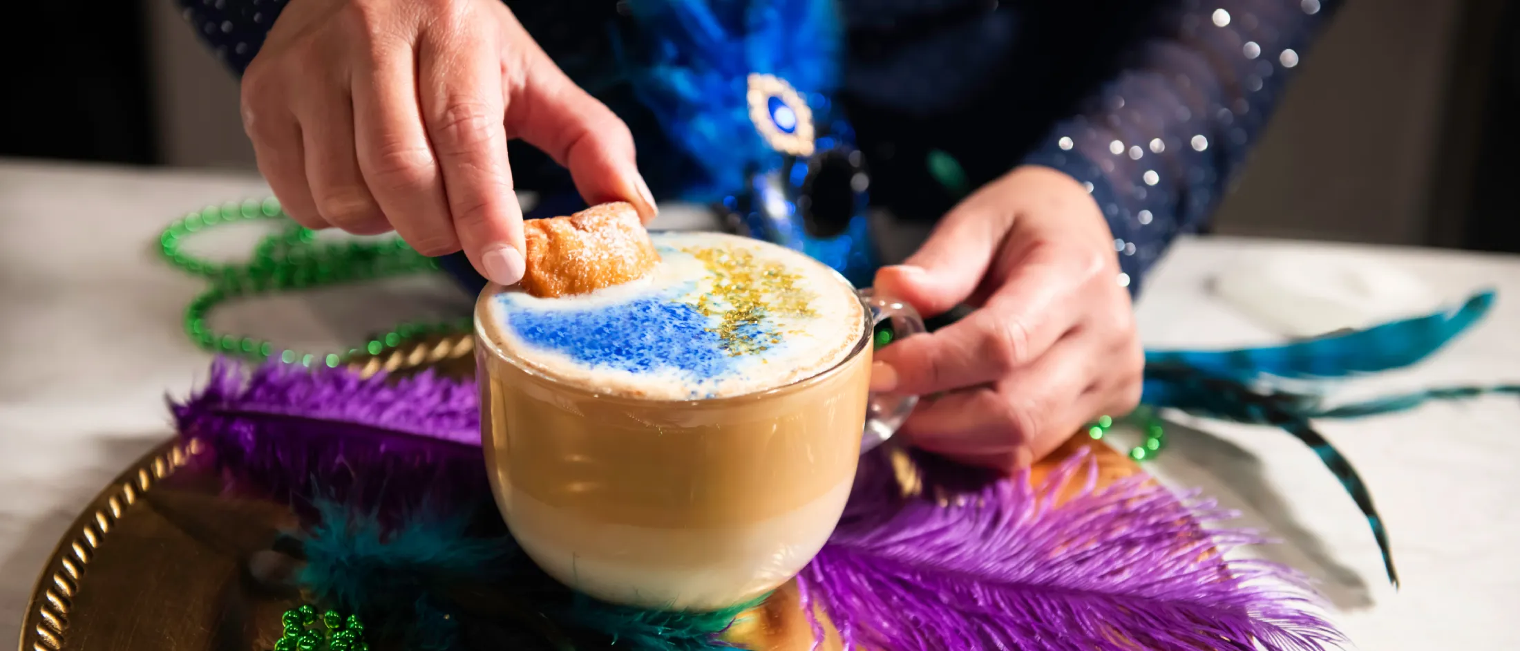 Person dipping a small pastry into a latte with blue and gold foam art on a tray decorated with colorful feathers and beads.