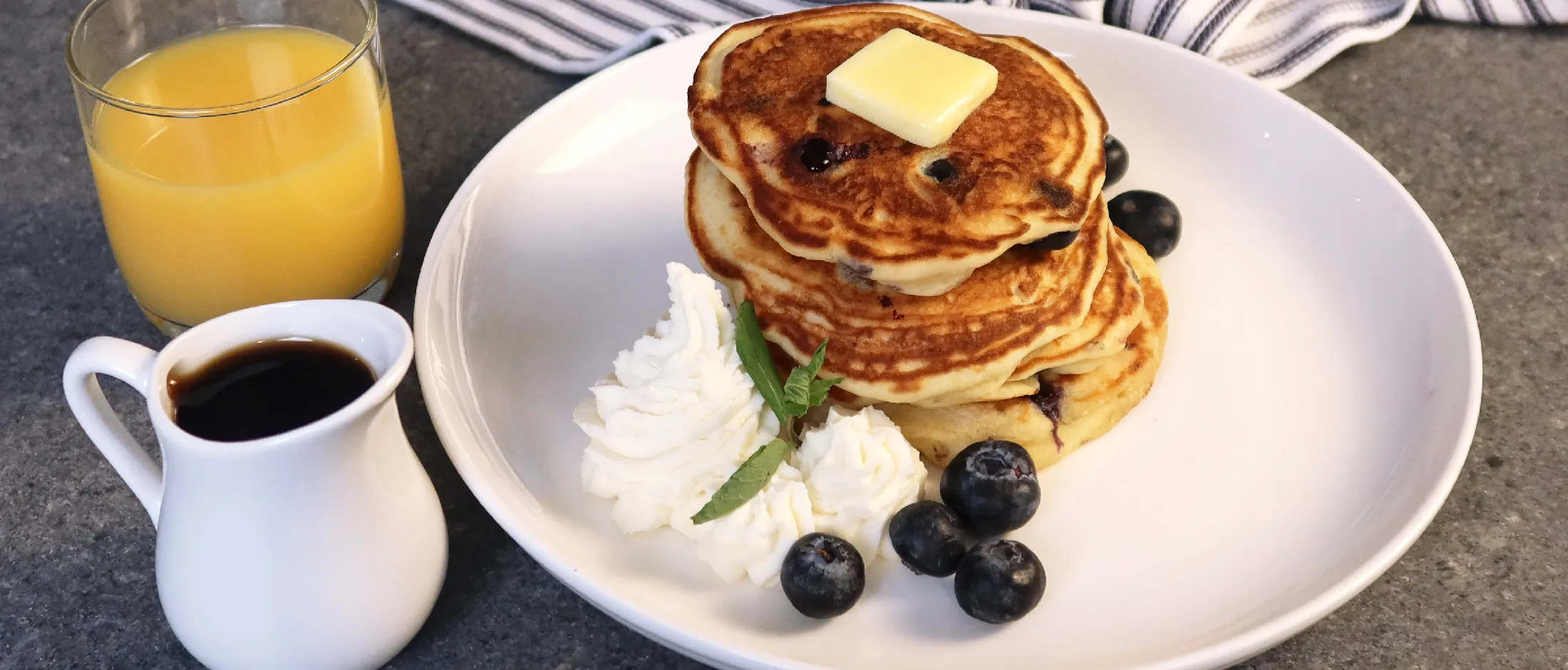 Stack of blueberry pancakes with butter, whipped cream, and fresh blueberries on a white plate, next to orange juice and syrup.