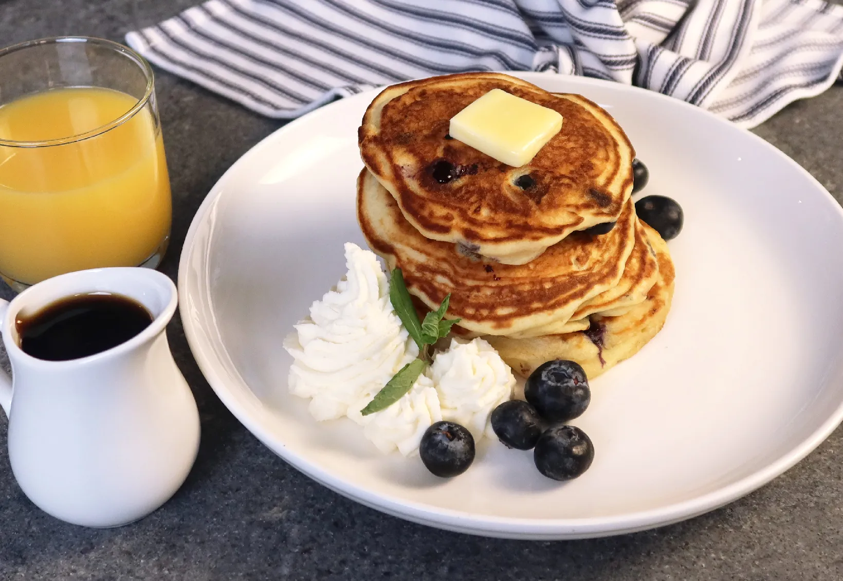 Stack of pancakes with butter, whipped cream, and blueberries on a white plate, served with orange juice and syrup in a white pitcher.