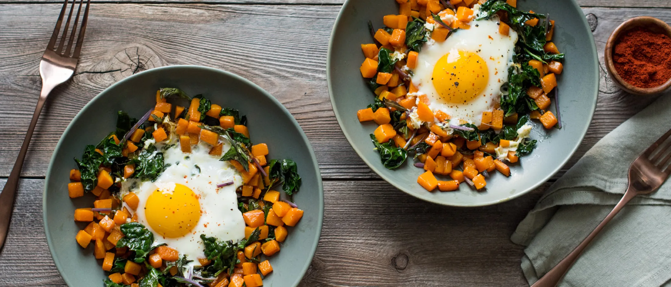 Two plates with sunny-side-up eggs surrounded by sautéed sweet potatoes and spinach on a wooden table with forks and a spice bowl.