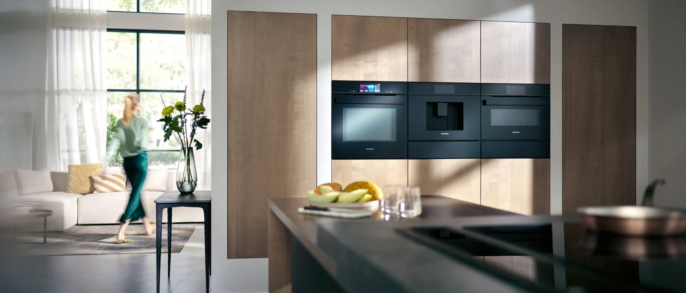 Cucina moderna con forni a muro, un'isola cucina e una persona sfocata che cammina vicino a un luminoso soggiorno con grandi finestre.