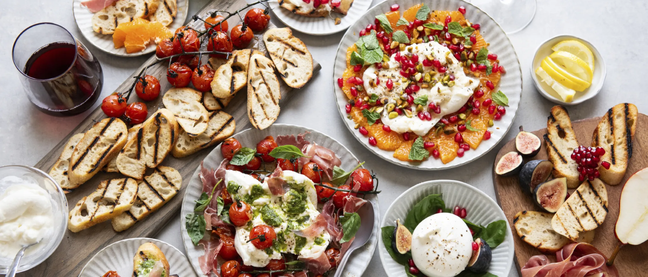 Assorted grilled bread, burrata cheese, roasted tomatoes, figs, pomegranate seeds, greens, prosciutto, and drinks on a light table.