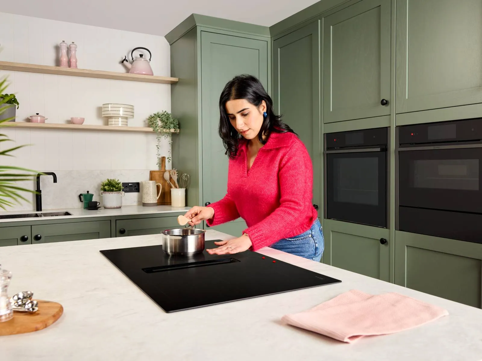 A woman who is visually impaired, wearing a pink sweater leans over a saucepan on an induction hob, stirring with a wooden spoon.