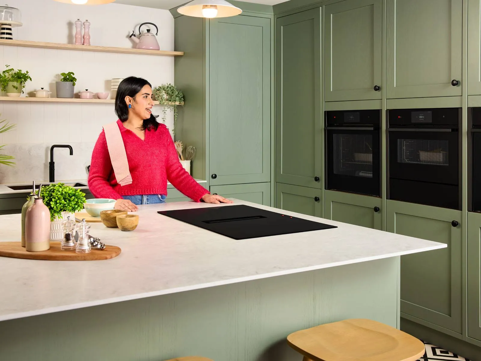 Visually impaired women in pink sweater with tea towel over her shoulder stands at a marble island in a sage-green kitchen with ovens and hob using voice command feature on smart device.