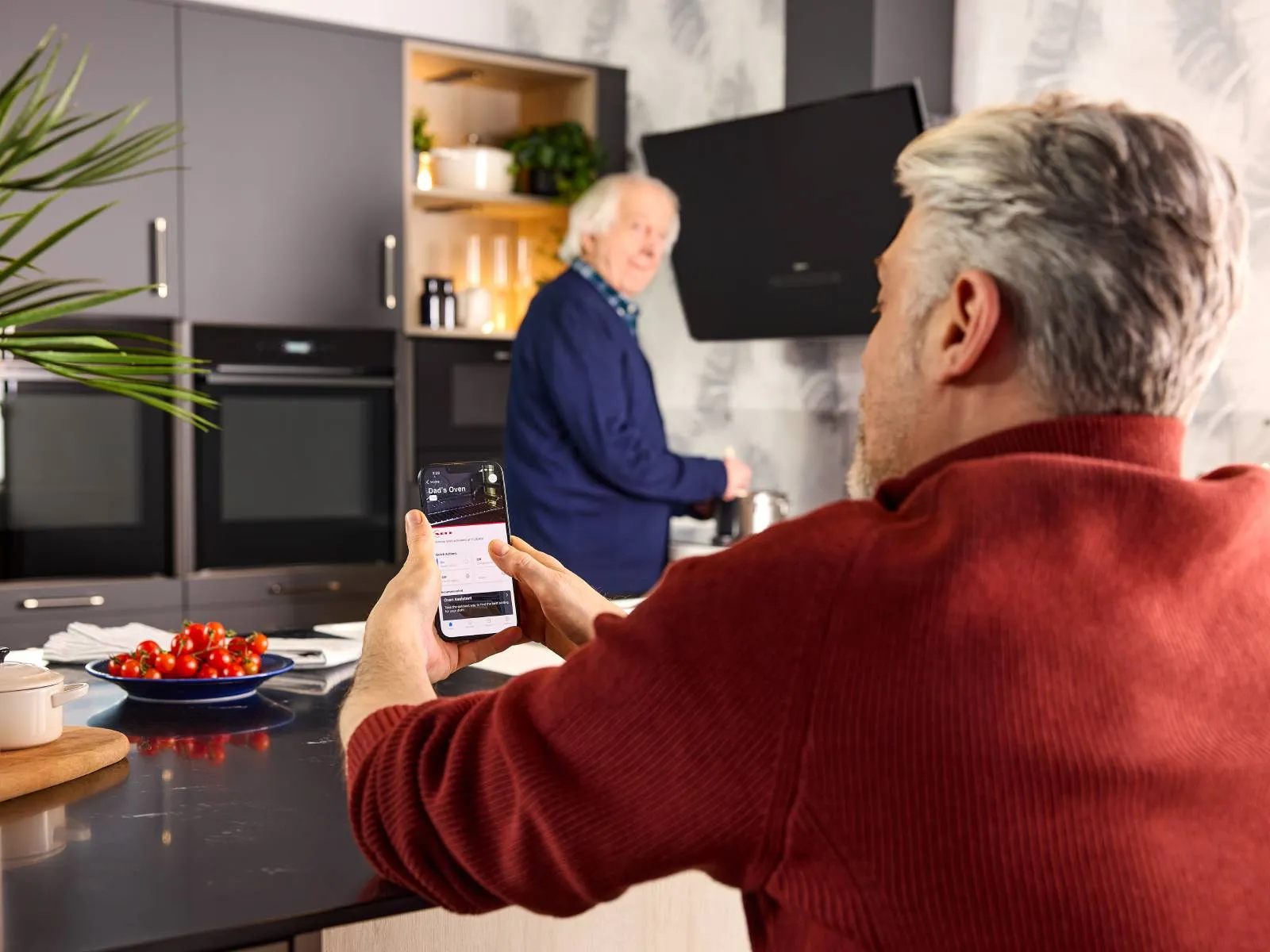 An elderly person cooks at the hob. His son monitors the Home Connect app remotely for added safety and peace of mind.