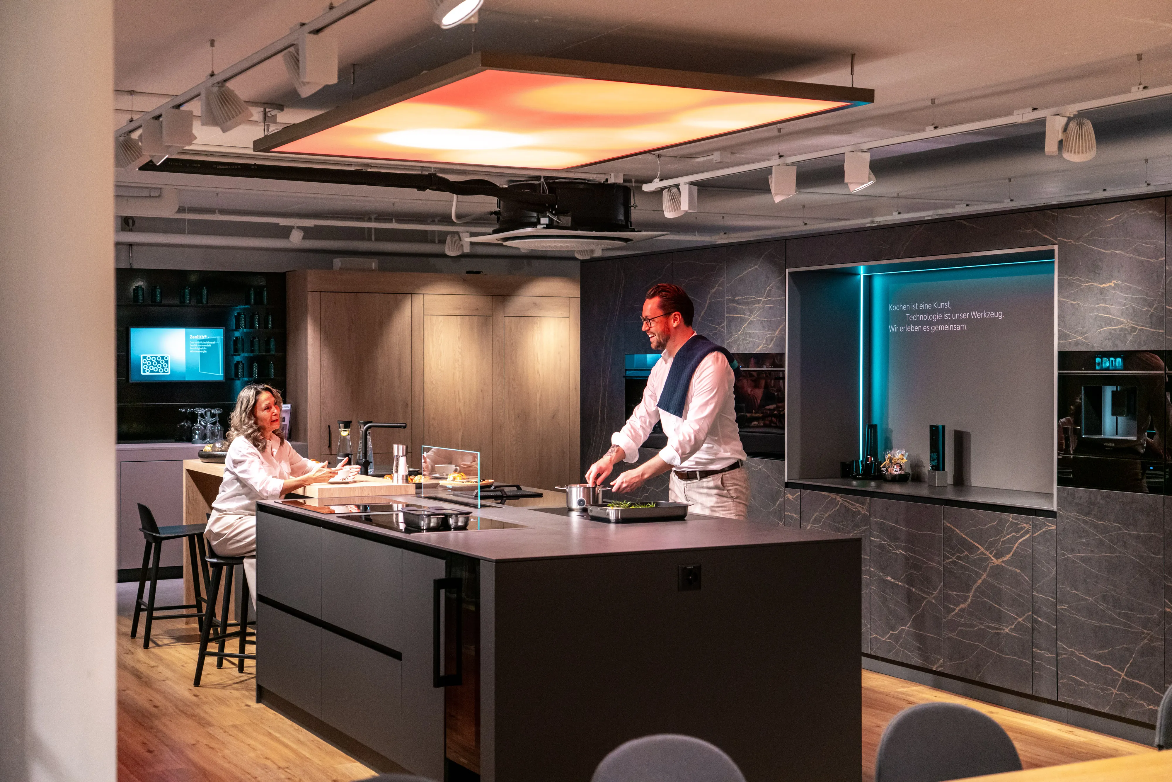 Kochvorführung in einem modernen Siemens Showroom: Ein Berater kocht für eine Kundin an einer dunklen Kücheninsel mit Einbaugeräten.