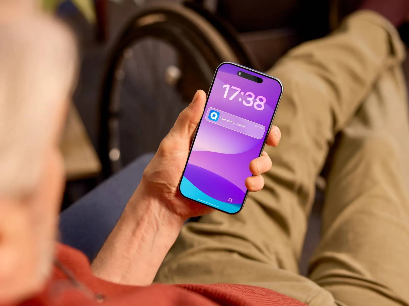 Close-up of a hand holding a smartphone showing a purple lock screen with the time 17:38 and a notification that reads “Your dish is ready,” with a blurred wheelchair wheel in the background.