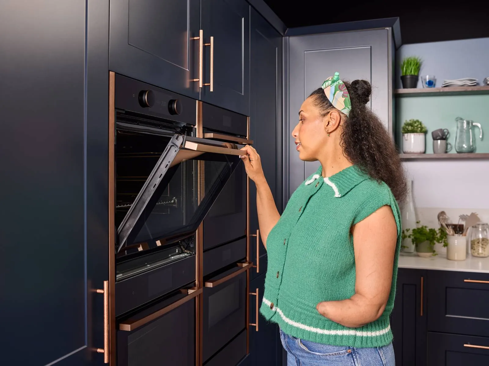 Woman with a limb difference affecting one forearm, opening the slide and hide oven door with one hand, standing beside navy cabinetry and open shelving with plants and dishes behind.