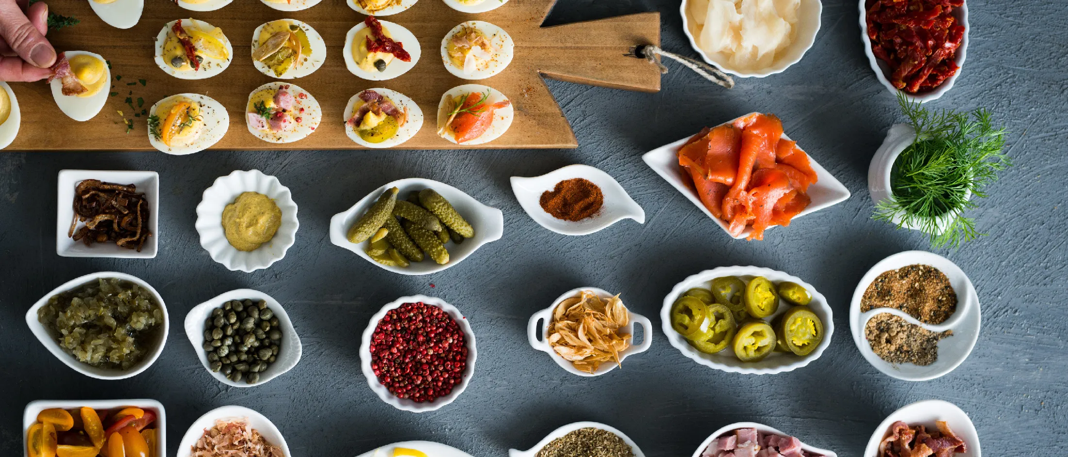 Assorted deviled eggs topped with various garnishes, surrounded by small bowls of pickles, spices, smoked salmon, and fresh herbs on a dark surface