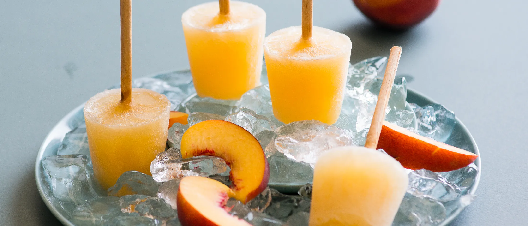 Four peach popsicles on wooden sticks served on ice with fresh peach slices on a blue plate