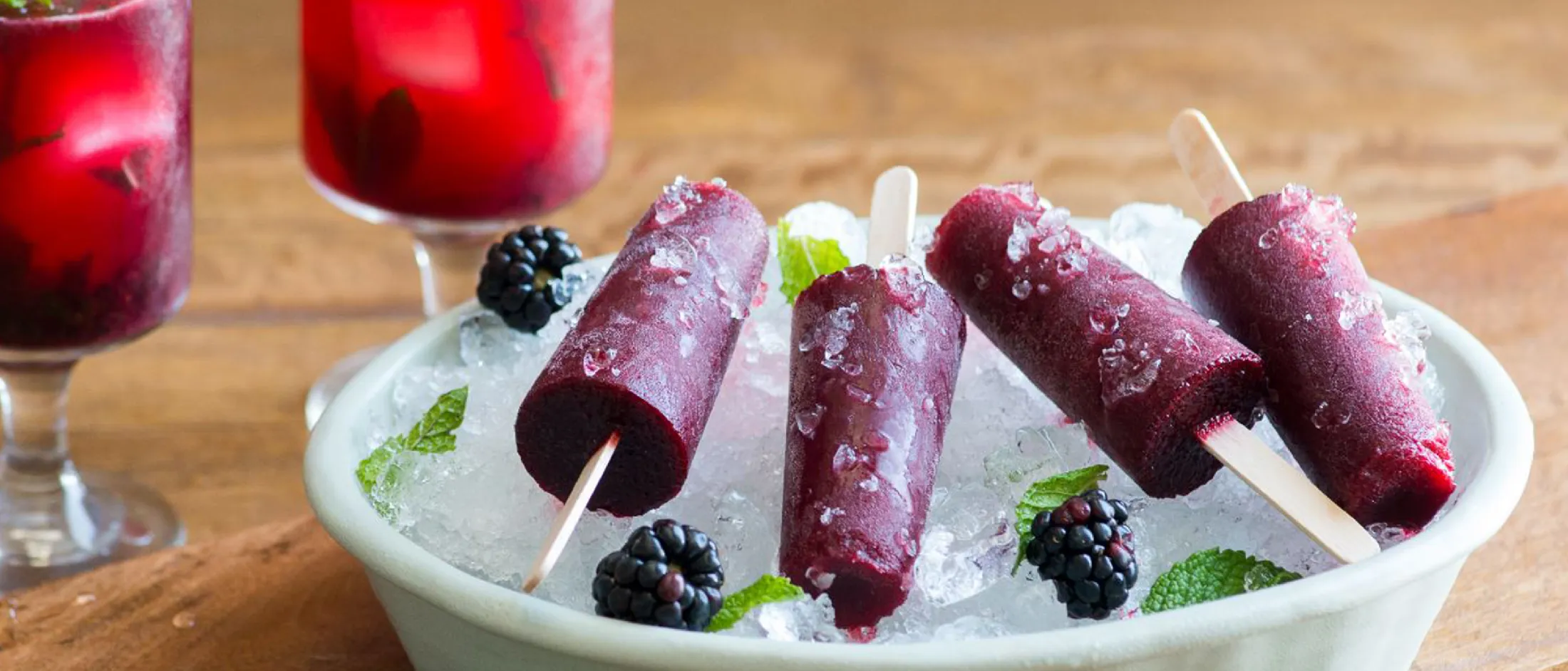 Frozen berry popsicles in a bowl, surrounded by mint leaves and blackberries, with red berry drinks in the background