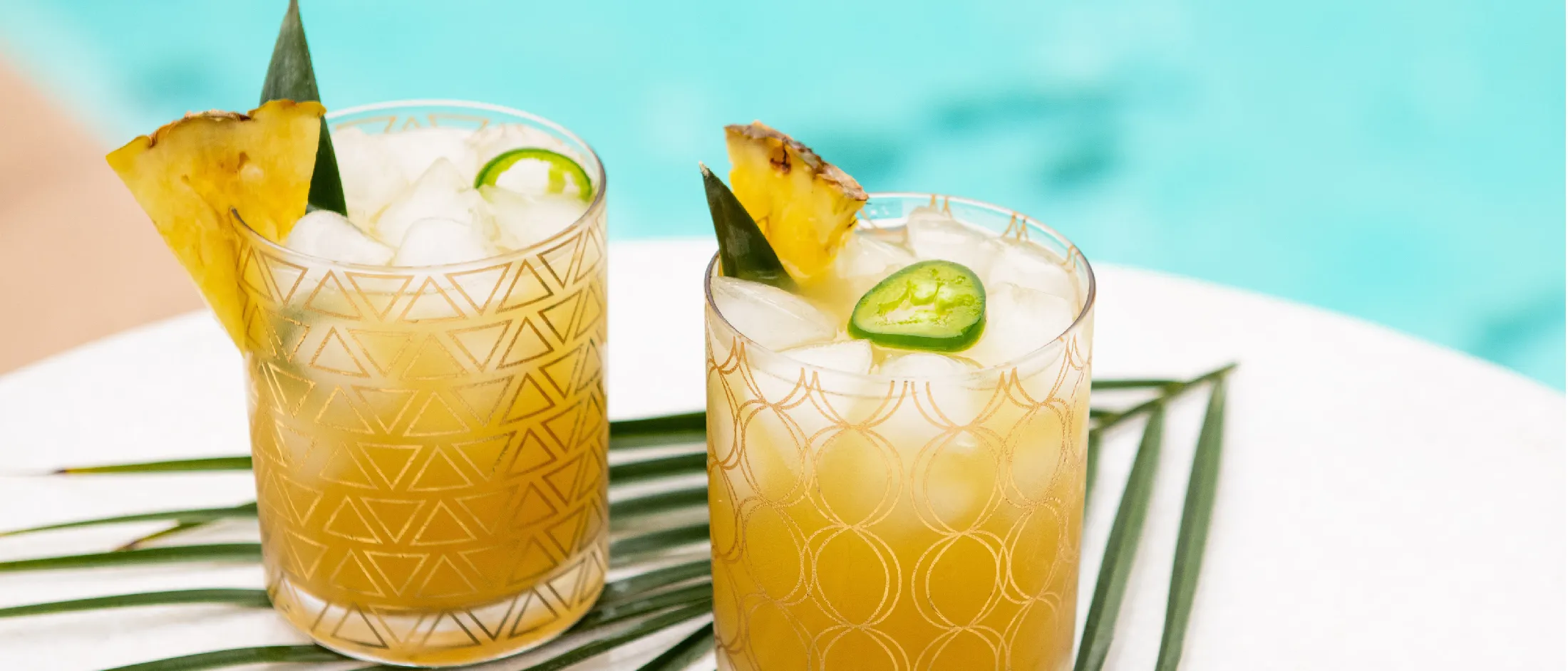 Two iced tropical cocktails garnished with pineapple wedges and jalapeño slices, set on a white table by a bright blue pool.