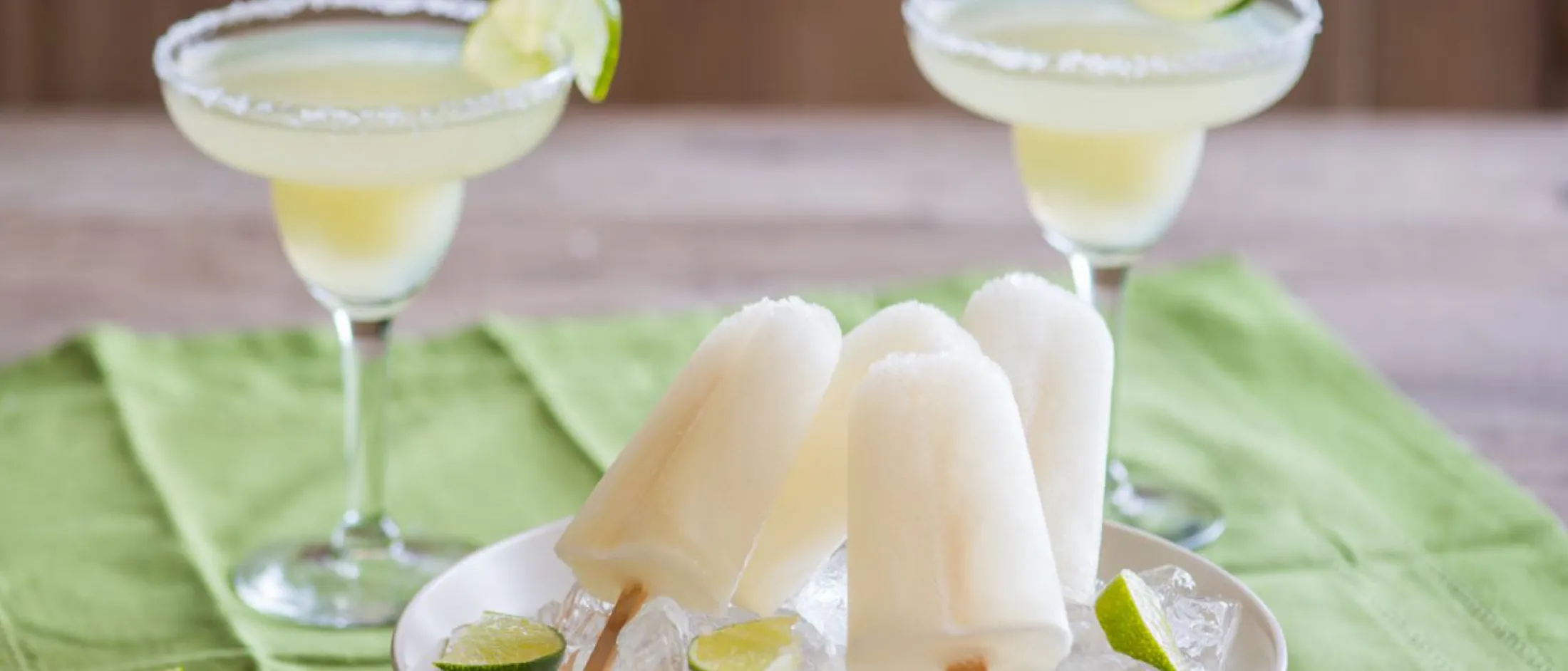 Margarita glasses, lime slices, and popsicles on a wooden surface with a green cloth