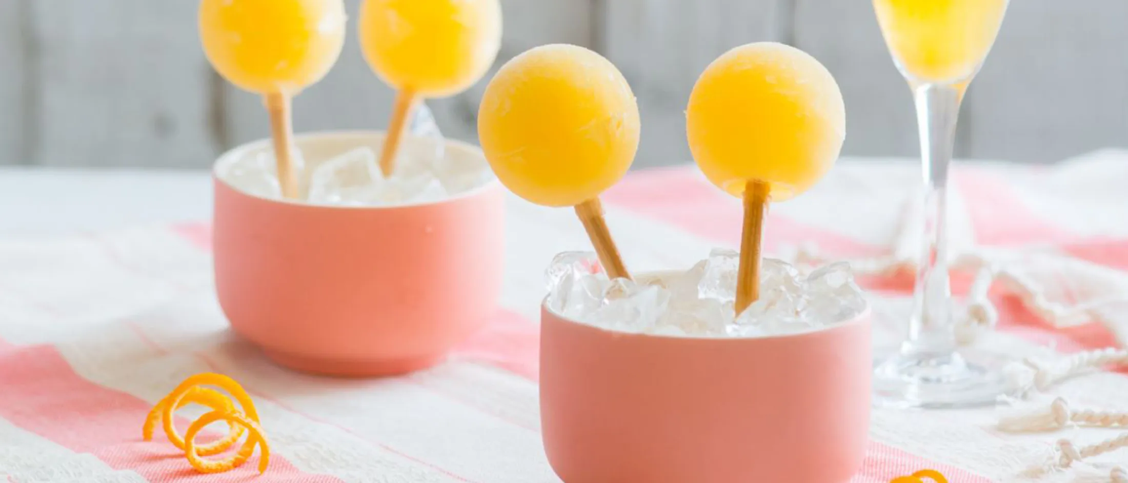Two pink cups filled with ice holding yellow frozen spherical pops on sticks, a glass of yellow drink, and orange peel twists on a table